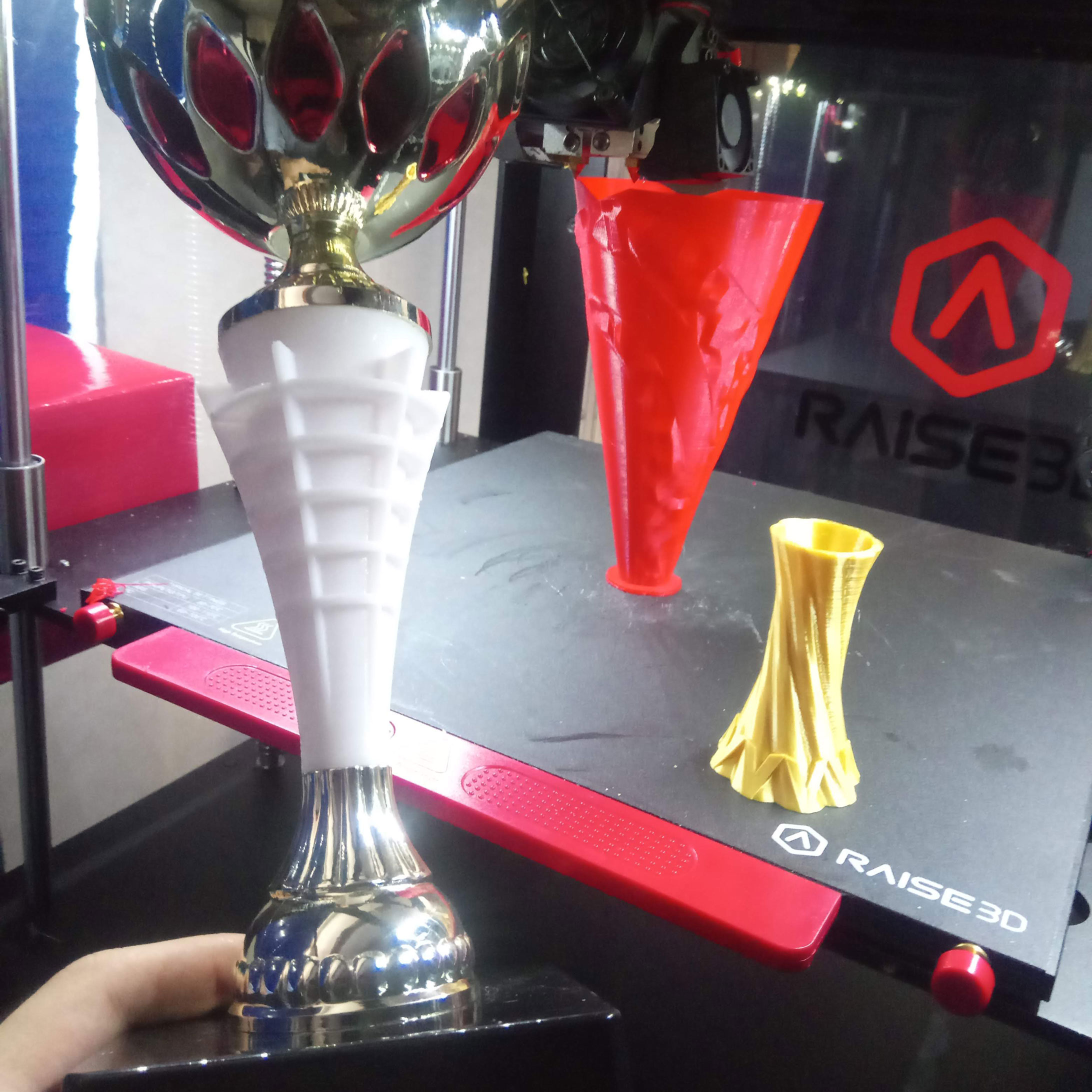 Une coupe tenue devant une imprimante 3D et une pièce rouge en cours de fabrication.
